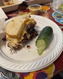 Pastrami sandwich on rye bread with melted cheese served with a whole pickle on a white plate