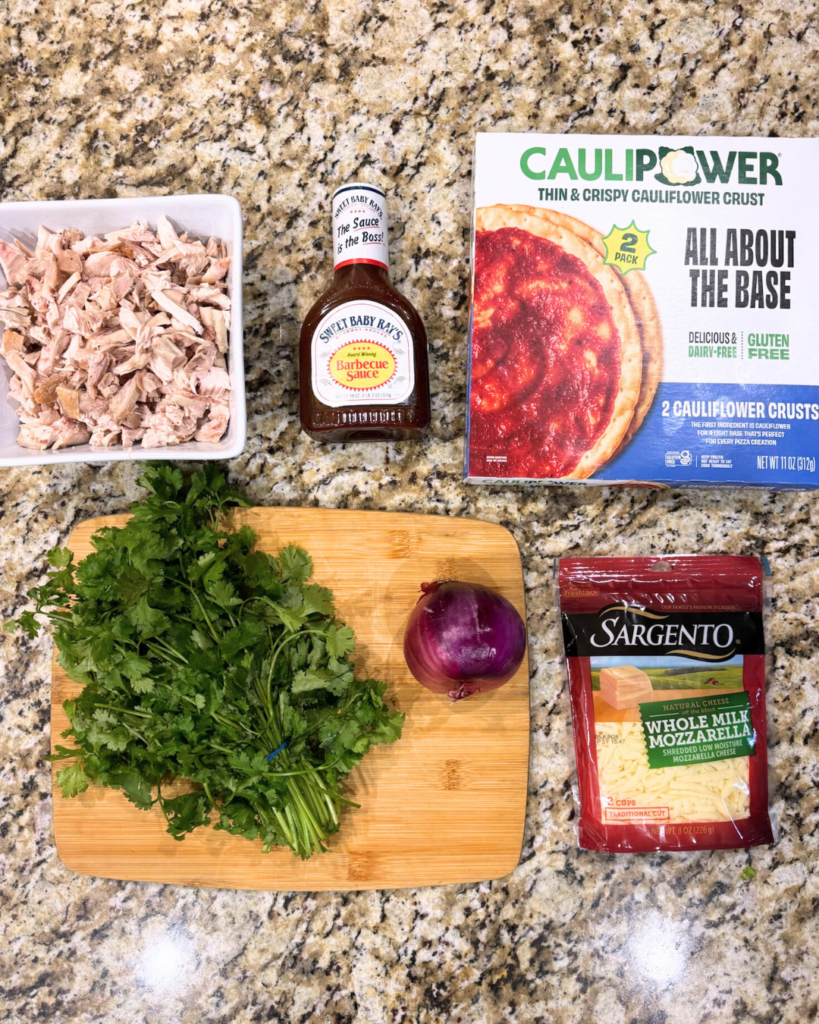 Flat lay of ingredients for BBQ chicken pizza on a granite countertop, including chopped rotisserie chicken, barbecue sauce, cauliflower pizza crusts, shredded mozzarella cheese, fresh cilantro, and a whole red onion on a wooden cutting board.