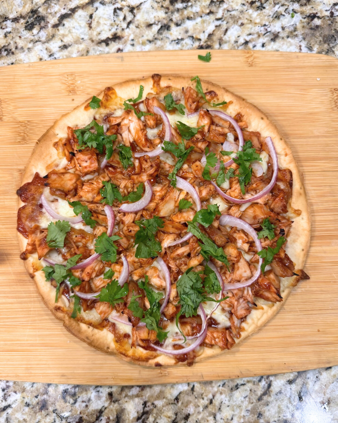 Overhead view of a homemade BBQ chicken pizza on a wooden cutting board, topped with shredded barbecue chicken, melted mozzarella cheese, sliced red onion, and fresh chopped cilantro on a golden baked crust