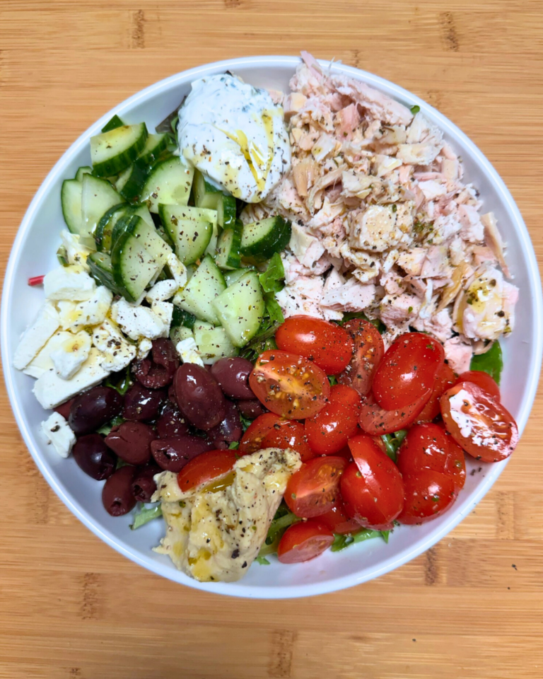 Greek chicken bowl with rotisserie chicken, vegetables, feta, hummus, and tzatziki in a white bowl.