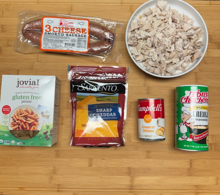 Ingredients for Cajun chicken and sausage pasta including rotisserie chicken, smoked sausage, gluten-free penne pasta, shredded cheddar cheese, cream of chicken soup, and Cajun seasoning arranged on a wooden cutting board.