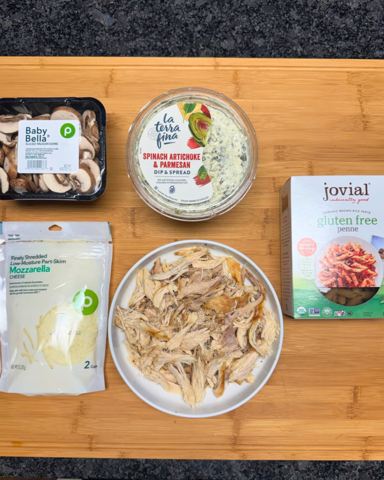 Ingredients for spinach artichoke chicken pasta bake with rotisserie chicken, mushrooms, mozzarella, penne pasta, and spinach artichoke dip on a wooden board.