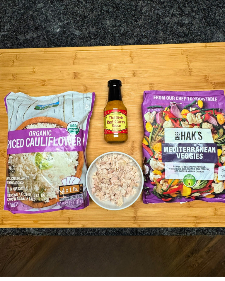 Ingredients for Thai chicken curry with rotisserie chicken, including cauliflower rice, frozen Mediterranean vegetables, Trader Joe’s red curry sauce, and chopped chicken on a cutting board.