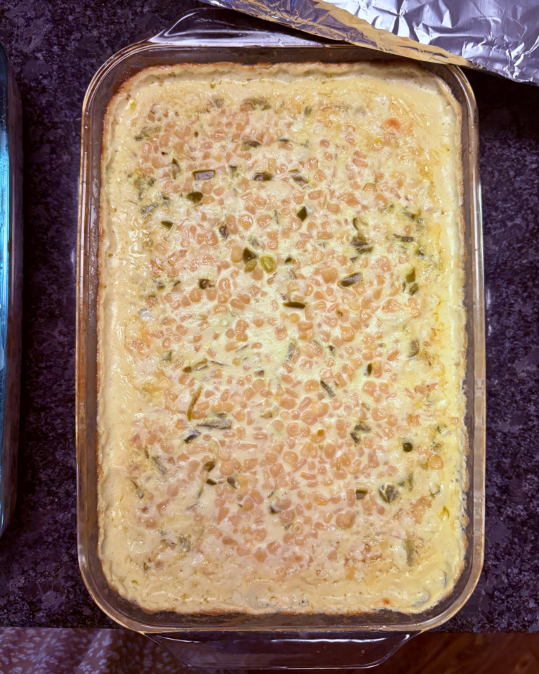 A freshly baked corn casserole in a rectangular glass dish, with a golden, creamy top and visible corn kernels, sitting on a dark countertop with foil partially peeled back.
